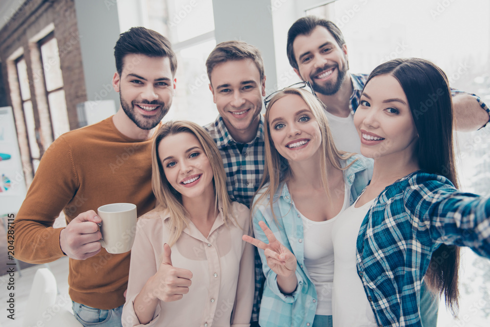 Fototapeta premium Self portrait of cheerful, joyful, positive students managers shooting selfie on front camera in modern office showing two fingers, like, peace, hi sign, holding cup with coffee, partnership