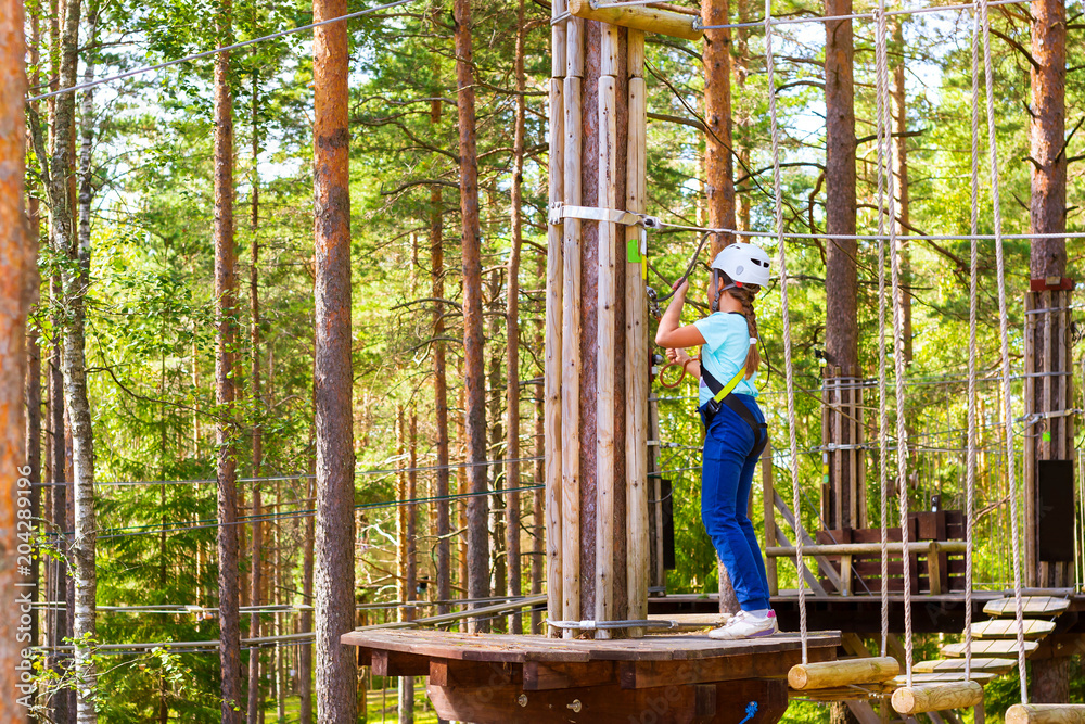 Teenage girl goes on hinged trail in extreme rope Park in summer forest. High-altitude climbing training of child on adventure track, equipped with safety straps and protective helmet. Estonian summer Teenage girl goes on hinged trail in extreme rope Park in summer forest. High-altitude climbing training of child on adventure track, equipped with safety straps and protective helmet. Estonian summer