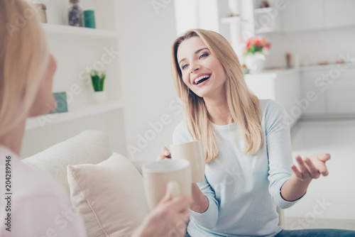 Portrait of laughing cheerful positive girl joking sharing secrets with her mother having mugs with tea cacao beverage in hands cozy comfort atmosphere enjoying meeting indoor sitting on sofa