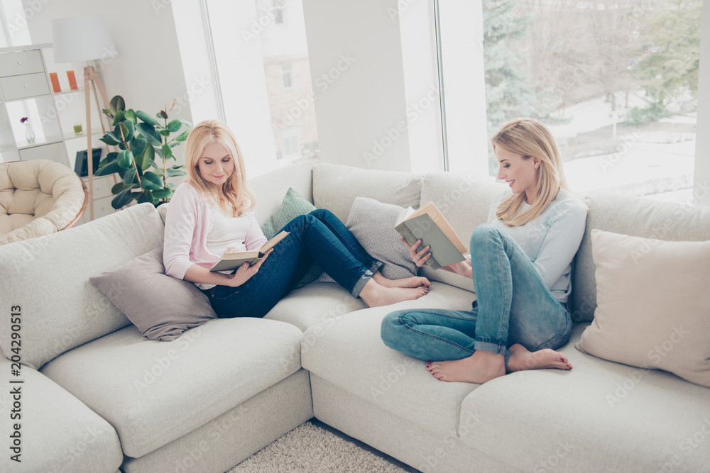 Portrait of attractive clever mother and daughter sitting with legs on sofa holding books in hands detective lover enjoying free time in comfortable cozy atmosphere modern white apartments