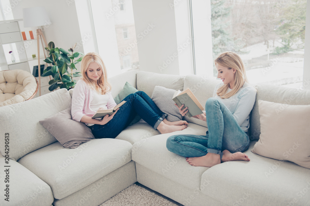 Portrait of attractive clever mother and daughter sitting with legs on sofa holding books in hands detective lover enjoying free time in comfortable cozy atmosphere modern white apartments Portrait of attractive clever mother and daughter sitting with legs on sofa holding books in hands detective lover enjoying free time in comfortable cozy atmosphere modern white apartments