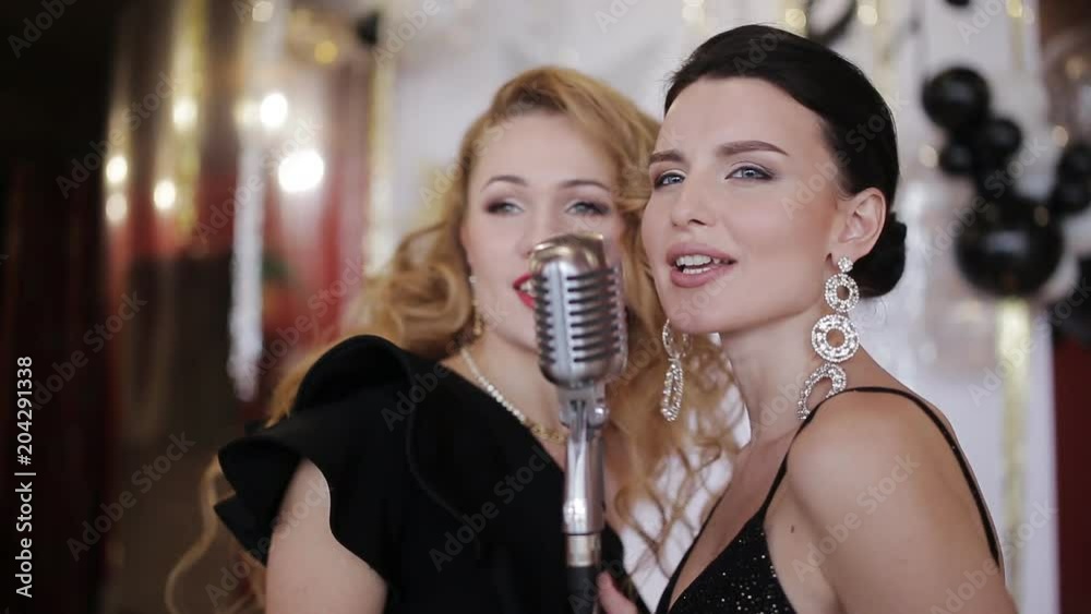 Two glamorous girls in evening gowns singing into a vintage microphone and having fun at the party Two glamorous girls in evening gowns singing into a vintage microphone and having fun at the party