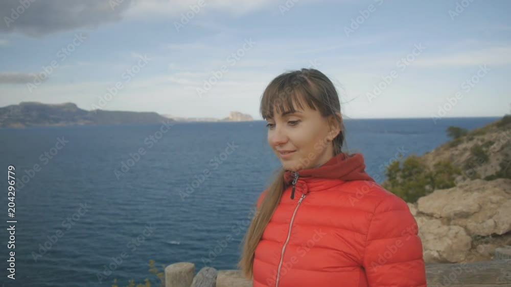 Young woman on sea background. Smiling caucasian brown-haired female in red hoodie and bubble jacket watching the beach from the observation deck Young woman on sea background. Smiling caucasian brown-haired female in red hoodie and bubble jacket watching the beach from the observation deck