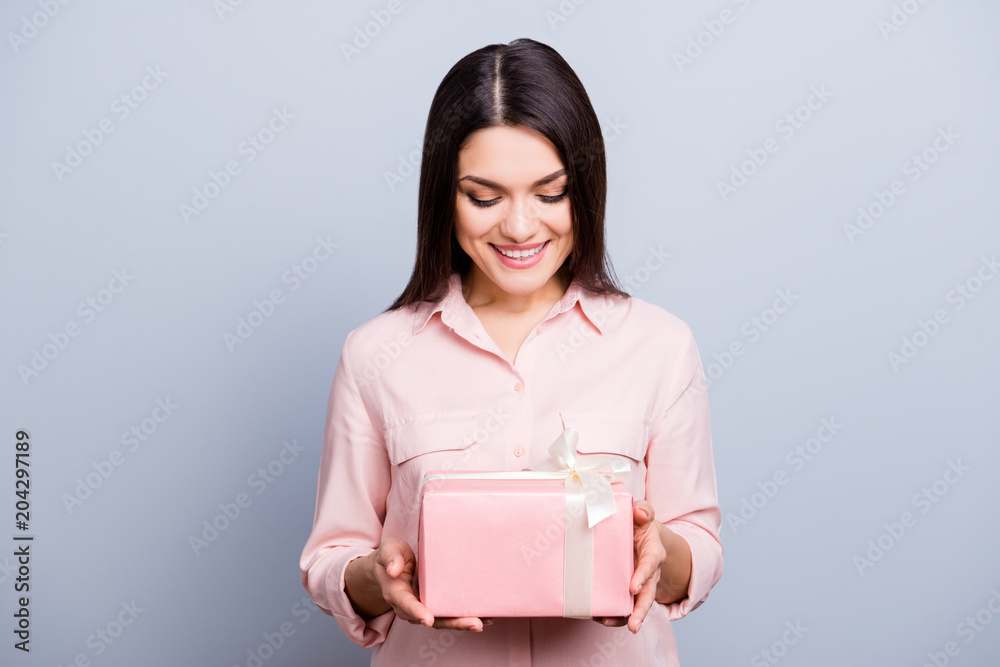 Portrait of glad, charming pretty, trendy woman having gift box in pink package with white bow looking at present isolated on grey background Portrait of glad, charming pretty, trendy woman having gift box in pink package with white bow looking at present isolated on grey background