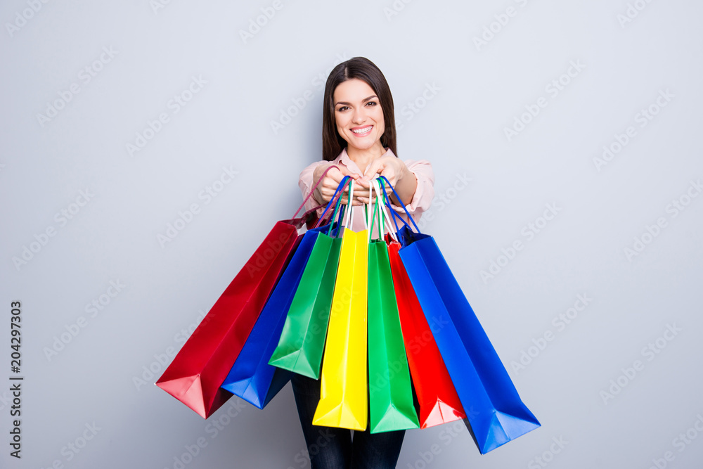 Portrait of pretty, charming, glad, positive woman giving many colorful packets front to the camera isolated on grey background, I bought for you these all gifts