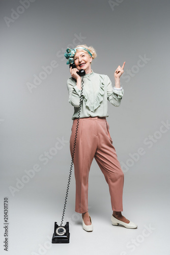 smiling senior woman talking by vintage phone and pointing up on grey