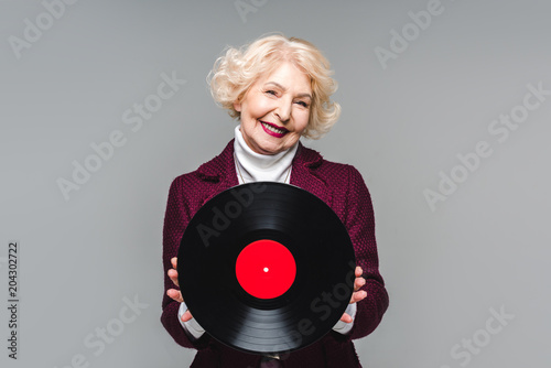 stylish senior woman holding vinyl disc isolated on gray background