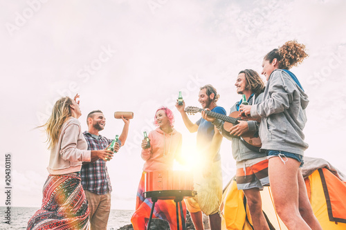 Group of friends having barbecue drinking beers while camping on the beach - Happy people enjoying camp bbq playing guitar, singing and listening music at sunset - Friendship, adventure concept