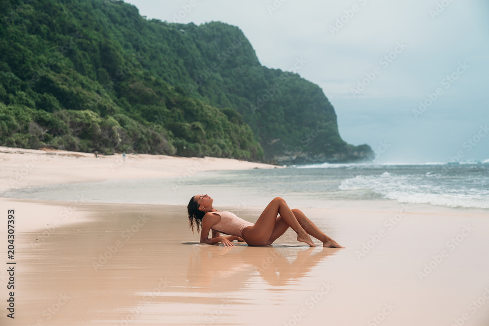 gorgeous young girl in a beige bikini lies on the beach, sunbathing near the ocean. A model with a sexy body and a sports figure in the clear sea water on the island. The concept of the weekend on