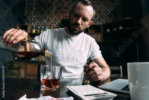 depressed young man pouring whiskey in glass on kitchen