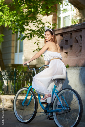 Smiling female in sunny day on a vintage bicycle at the city street looked around to look at the camera. The girl is dressed in a light dress and an accessory on her head