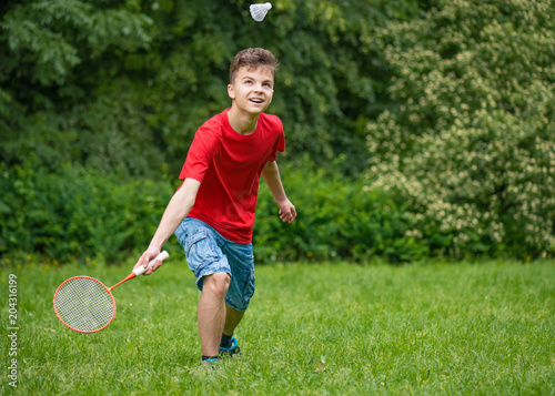 Young teen boy playing badminton in meadow with forest in background. Child with badminton rackets in hand. Kid have fun in summer park at day.