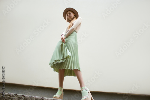 Young red hair girl in hat, dressed in beige blouse and turqoise pleats skirt, on light street backgraund.  Fashion and stylish concept.