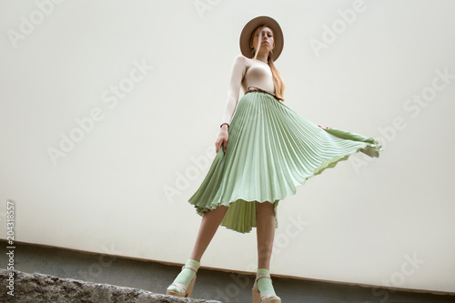 Young red hair girl in hat, dressed in beige blouse and turqoise pleats skirt, on light street backgraund.  Fashion and stylish concept.