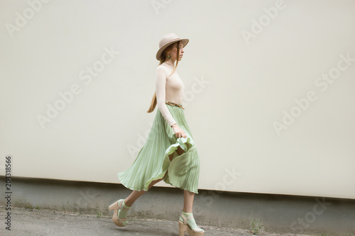 Young red hair girl in hat, dressed in beige blouse and turqoise pleats skirt, on light street backgraund.  Fashion and stylish concept.