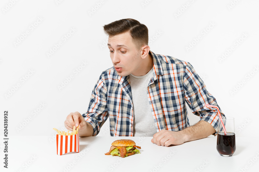 Handsome young man in shirt sitting at table with burger, french fries, cola in glass isolated on white background. Proper nutrition or American classic fast food. Advertising area with copy space. Handsome young man in shirt sitting at table with burger, french fries, cola in glass isolated on white background. Proper nutrition or American classic fast food. Advertising area with copy space.