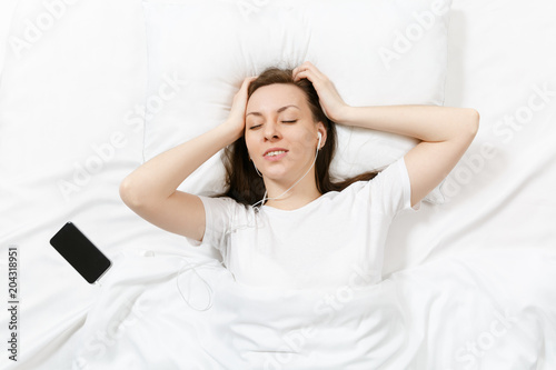 Top view of happy young woman lying in bed with white sheet, pillow, blanket, listen music from earphones in mobile phone. Calm beautiful female spending time in room. Rest, relax, good mood concept.