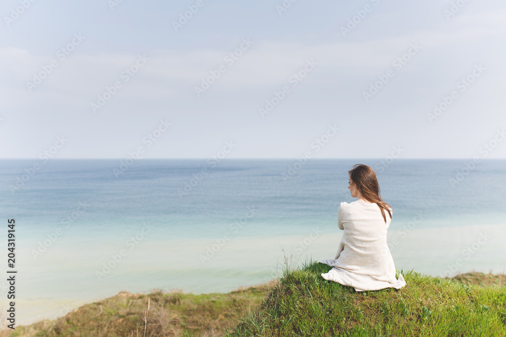 Young brunette relaxed beautiful woman in light casual clothes sitting back on grass looking aside in green field on water background. Beautiful nature landscape. Lifestyle, leisure concept.