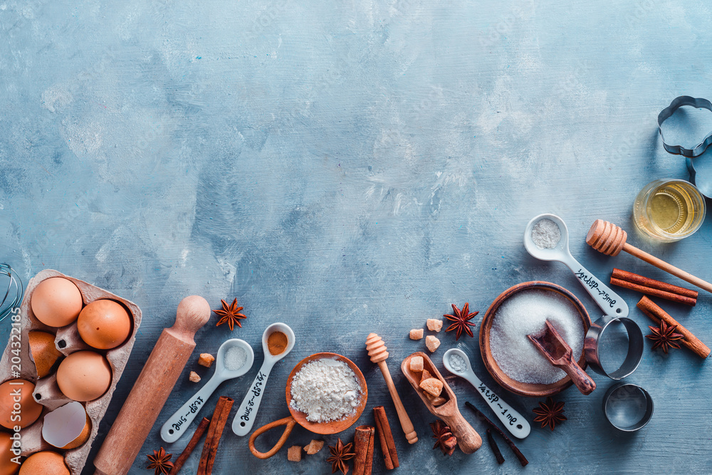 Cooking tools and ingredients flat lay with copy space. Baking header with measuring  spoons, wooden scoops, whisks, rolling pin, cookie cutters, sugar, flour,  eggs and cinnamon on a blue background. Stock Photo