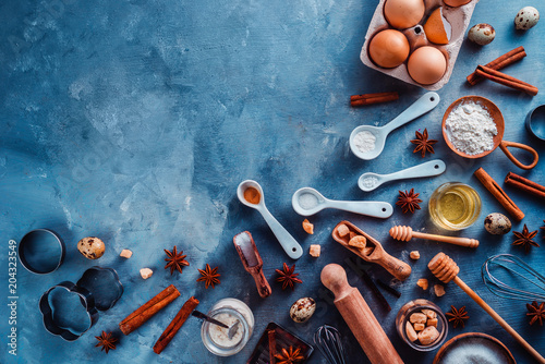 Cooking tools and ingredients flat lay with copy space. Baking header with measuring spoons, wooden scoops, whisks, rolling pin, cookie cutters, sugar, flour, eggs and cinnamon on a blue background.