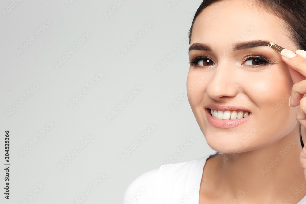 Young woman plucking eyebrow with tweezers on light background Young woman plucking eyebrow with tweezers on light background