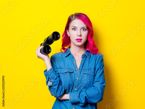 Young pink hair girl in blue shirt holding a binoculars. Portrait on isolated yellow background