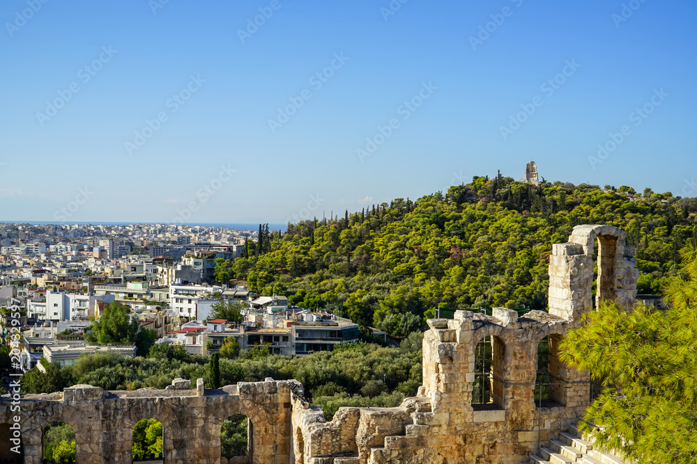 View of Athens cityscape and the Philopappu Monument through ancient stone theatre seeing lowrise white buildings architecture, mountain, trees and clear blue sky background