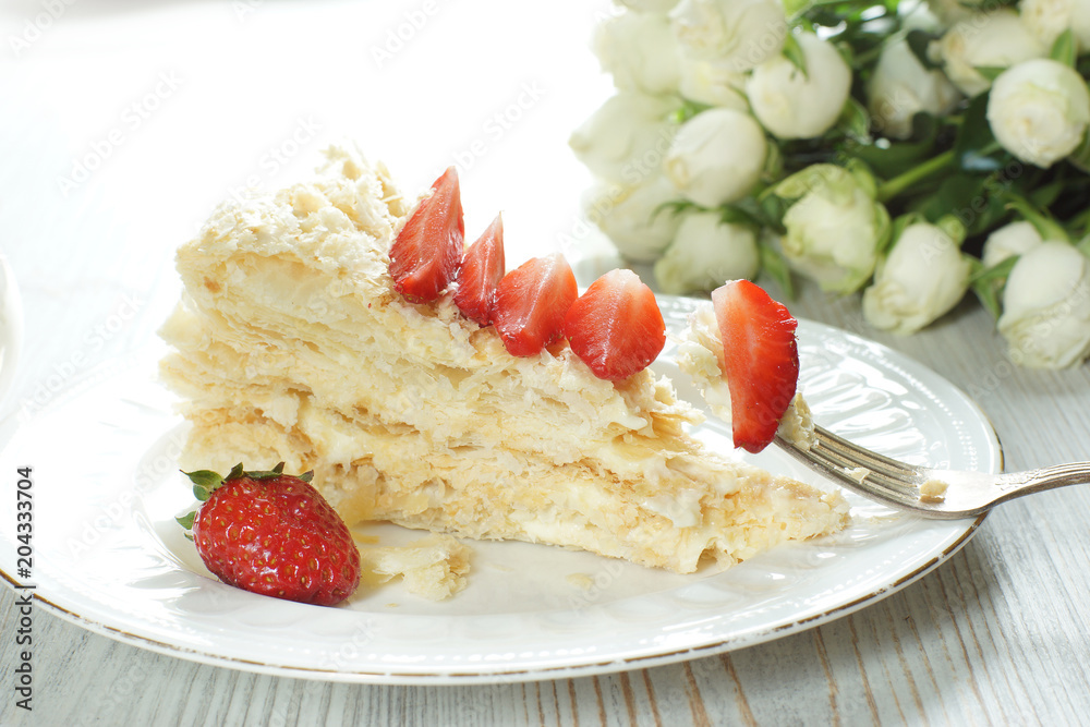 A piece of Napoleon cake on a plate adorned with a red ripe strawberry, a fork with a stuck piece of dessert A piece of Napoleon cake on a plate adorned with a red ripe strawberry, a fork with a stuck piece of dessert