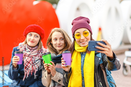 happy young women with take away cups outdoors taking selfie with smartphone