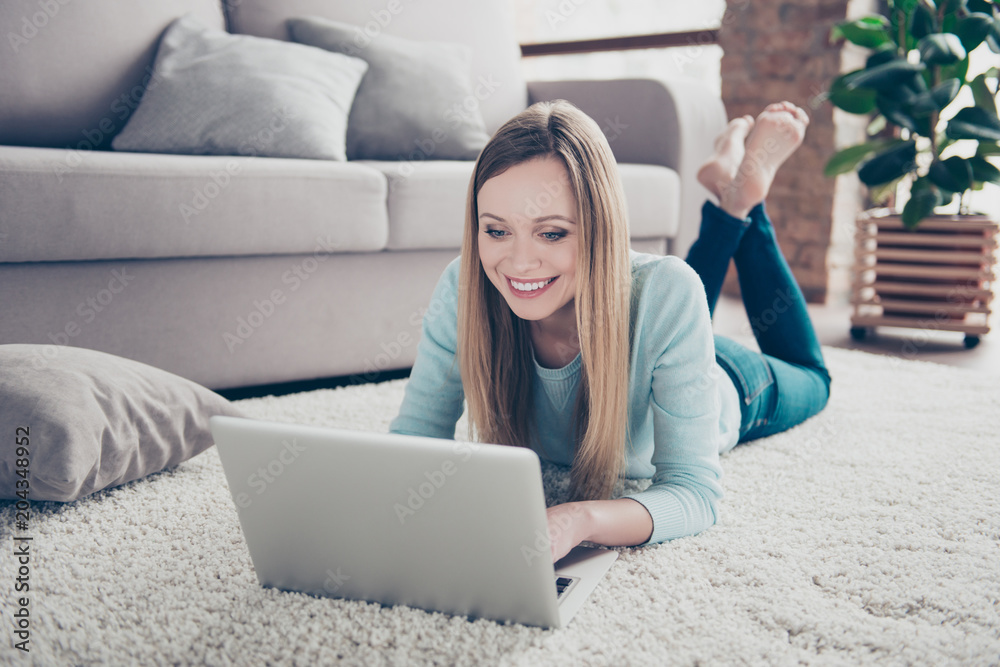Fototapeta premium Portrait of charming, pretty, blonde, domestic girl with hairstyle laying on the floor near sofa on stomach, checking email, using laptop and wifi internet, chatting with friends