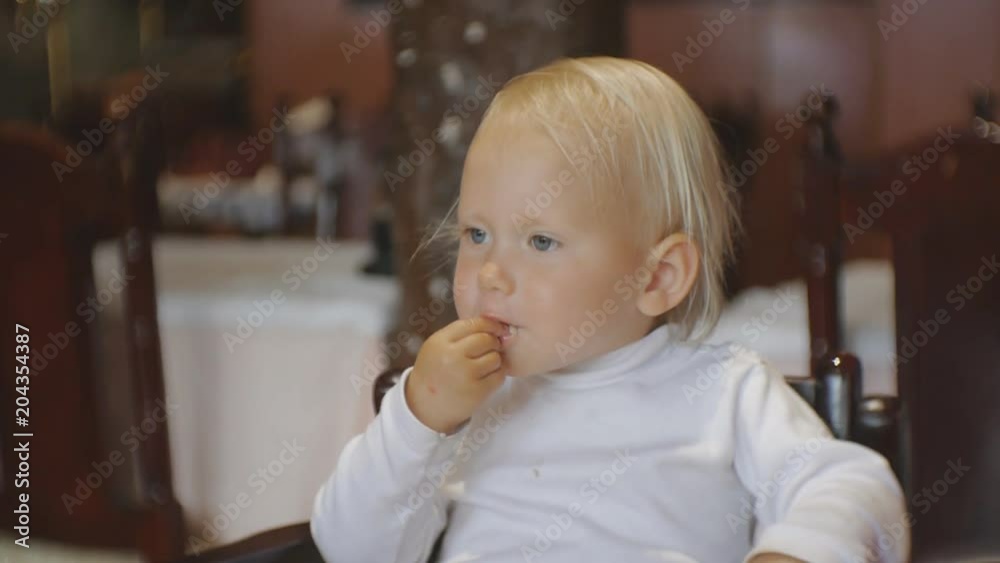 Baby eating food in restaurant. Blonde blue-eyed child in highchair takes pasta by hand from a plate putting it in the mouth Baby eating food in restaurant. Blonde blue-eyed child in highchair takes pasta by hand from a plate putting it in the mouth
