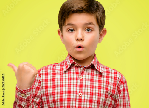 Handsome toddler child with green eyes happy and surprised cheering expressing wow gesture pointing up over yellow background