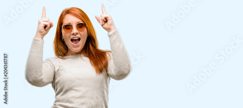 Beautiful young redhead woman happy and surprised cheering expressing wow gesture pointing up isolated over blue background