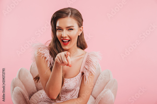 Obraz na plátně Portrait closeup of flirty winking woman in dress sitting in armchair and pointi
