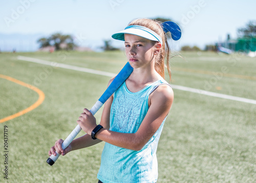 Sporty girl standing on sports field holding a blue hockey stick looking away.