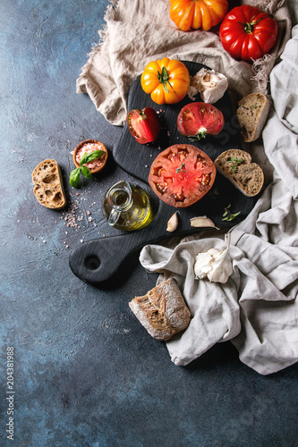 Variety of red and yellow organic tomatoes with olive oil, garlic, salt and bread for salad or bruschetta on wooden cutting board with linen cloth over blue texture background. Top view, space.