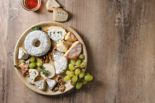 Cheese plate assortment of french cheese served with honey, walnuts, bread and grapes on ceramic plate over wood texture background. Top view, copy space.