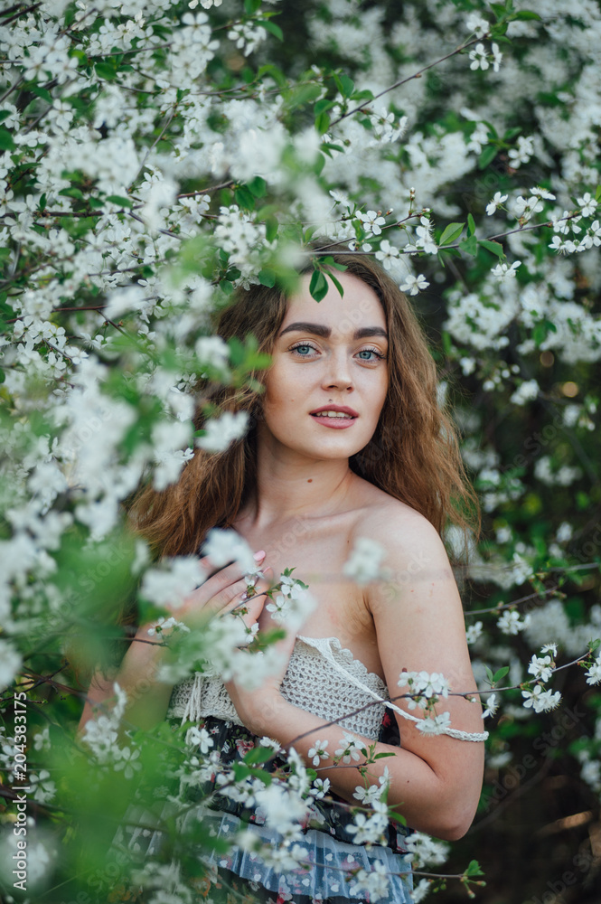 A beautiful girl in a dress stands next to a blooming cherry bush.