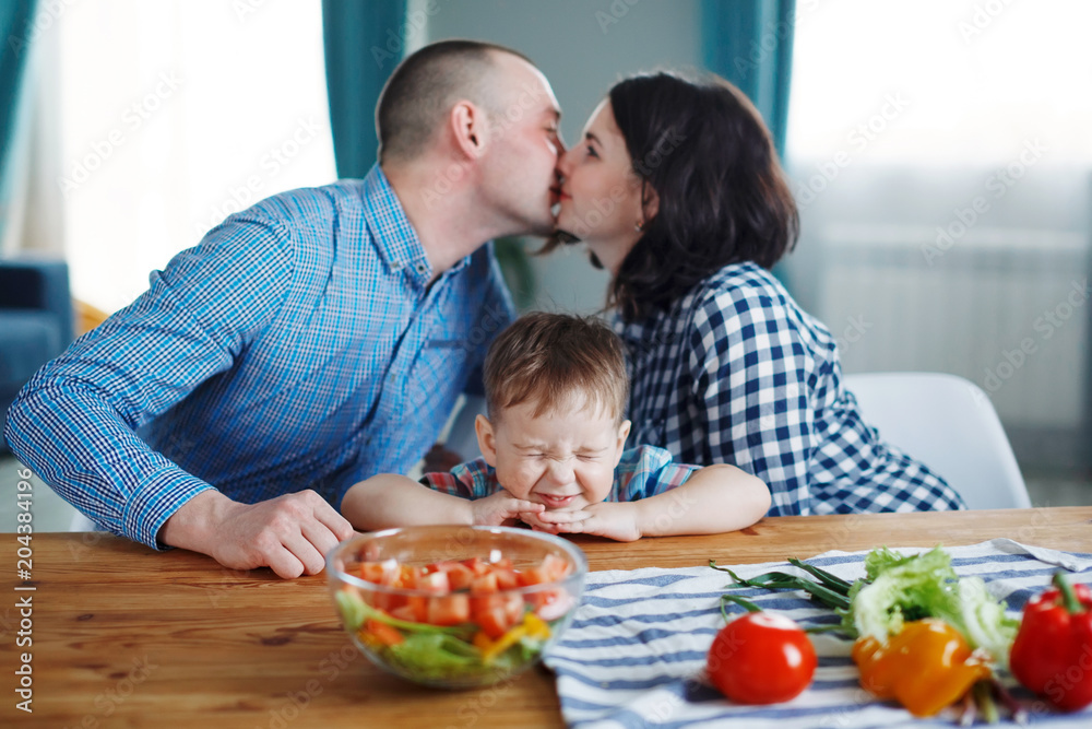 Young husband and wife kiss, son smiling, sitting at the table, family. Concept of a happy family healthy eating