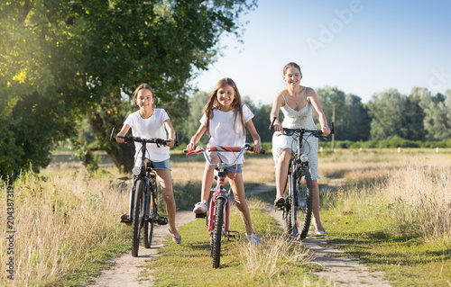 Two smiling girls riding bicycles with mother on countryside road