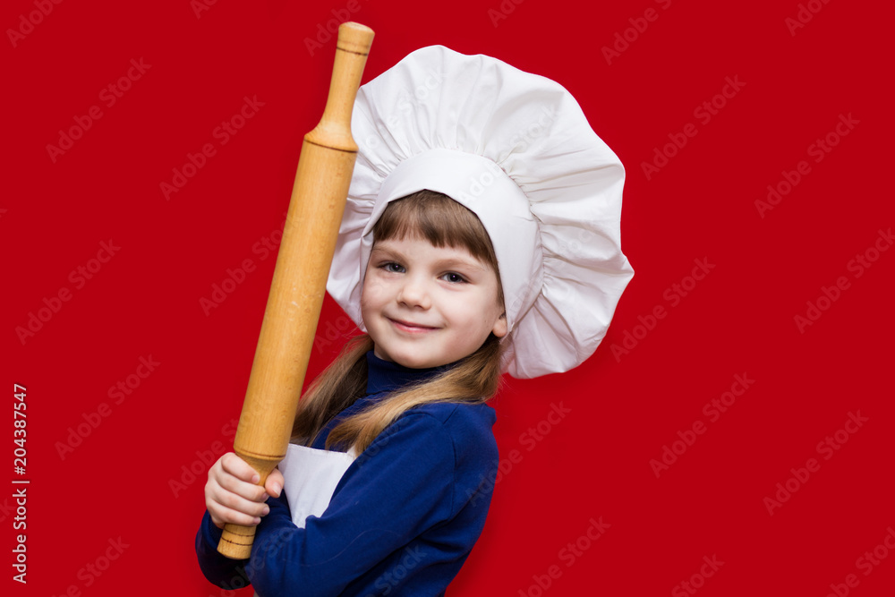 Happy little girl in chef uniform holds rolling pin isolated on red. Kid chef. Cooking Process Concept Happy little girl in chef uniform holds rolling pin isolated on red. Kid chef. Cooking Process Concept