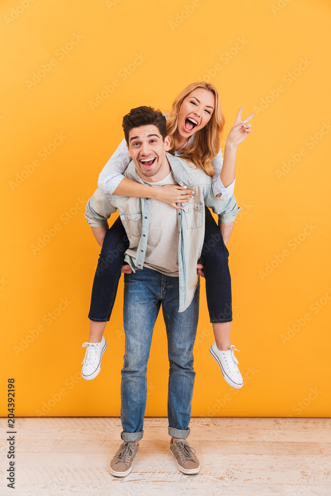 Full length photo of young couple having fun while guy giving piggyback ride to lovely girl showing peace sign, isolated over yellow background Full length photo of young couple having fun while guy giving piggyback ride to lovely girl showing peace sign, isolated over yellow background
