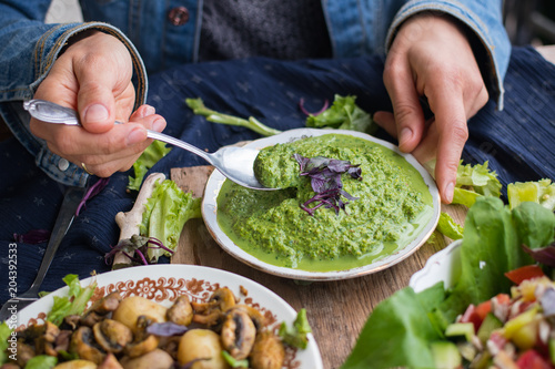 Woman hands holds avocado spinach green spread, dip Mix sauce with spoon. With basil leaves, ready for lunch or dinner with backed potato and vegetables salad. raw vegan vegetarian healthy food.