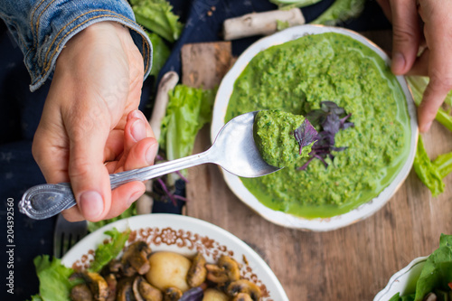 Woman hands holds avocado spinach green spread, dip Mix sauce with spoon. With basil leaves, ready for lunch or dinner with backed potato and vegetables salad. raw vegan vegetarian healthy food.