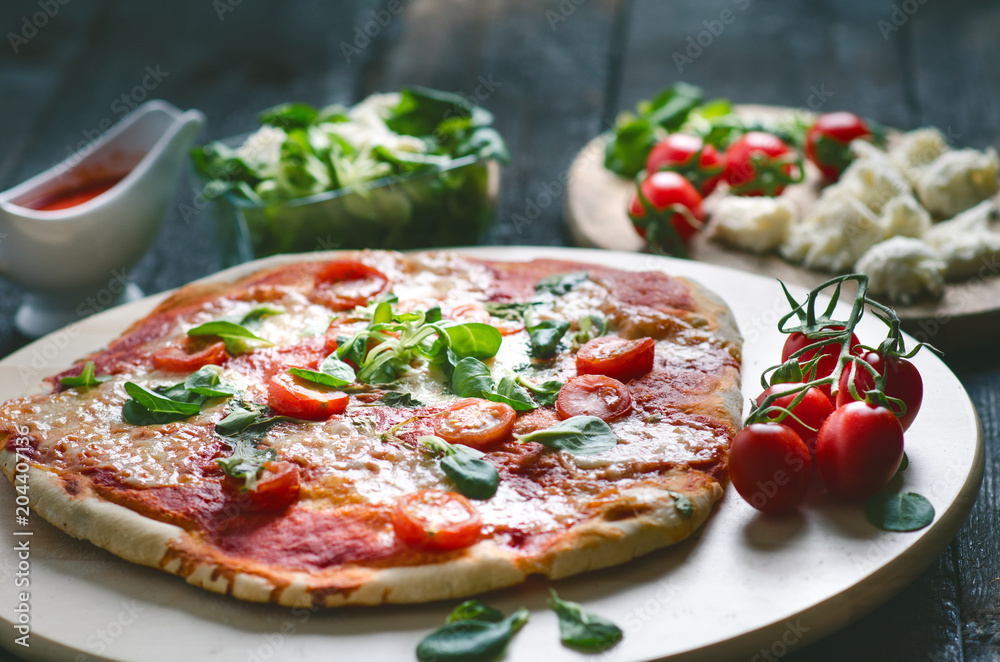 Italian food, cuisine. Margherita pizza on a black, wooden table with igredients like tomatoes, salad, cheese, mozzarella, basil. Delicious homemade food. 