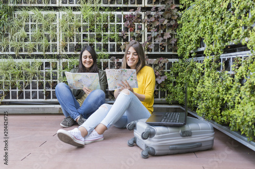 two beautiful women looking a map. green background. They are laughing and searching for a place to stay on holidays. Outdoors lifestyle and friendship. Suitcase and laptop besides.