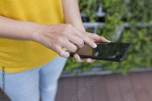 portrait outdoors of a beautiful young woman using on her mobile phone and smiling. Wearing a yellow casual shirt over green background. LIfestyle and fun.