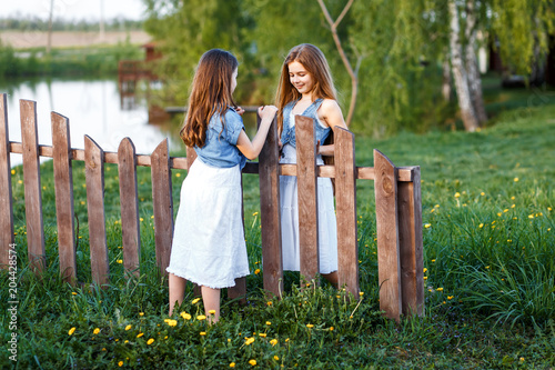 A portrait of two young girls with blue eyes in white dresses and jeans wests  in the garden near the fence enjoying, chattering and having fun ath the subset photo