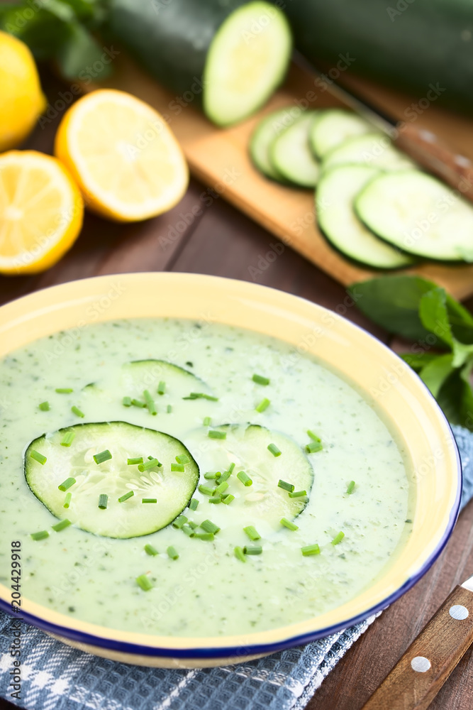 Refreshing cold cucumber, yogurt, mint and lemon soup in enamel bowl, garnished with cucumber slices and chives, ingredients in the back (Selective Focus, Focus one third into the image) Refreshing cold cucumber, yogurt, mint and lemon soup in enamel bowl, garnished with cucumber slices and chives, ingredients in the back (Selective Focus, Focus one third into the image)