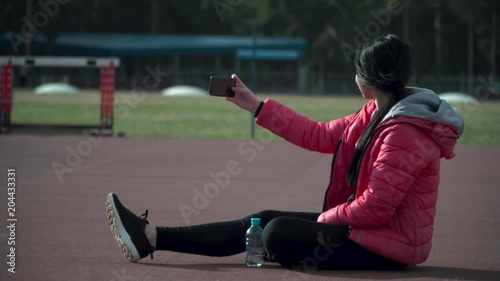 Brunette girl in a red jacket after training doing selfie on a smartphone sitting on the ground in the background of the stadium